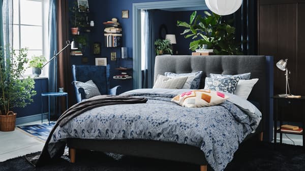A bedroom in different shades of blue with an upholstered bed, plenty of cushions, a wing chair, books and a large rug.