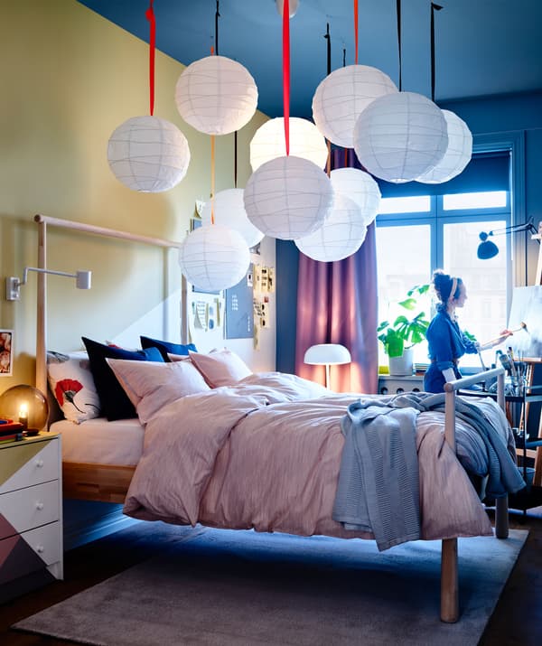 A bedroom styled with a pink colored SKOGSALM quilt cover and a grey colored throw on the bed. A woman in blue colored dress is paintaing near the window.