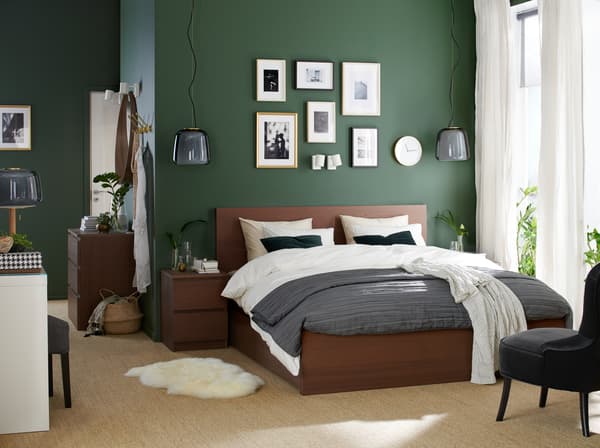 A bedroom with a bed frame and bedside table in brown-stained ash veneer, a white dressing table and a grey armchair.