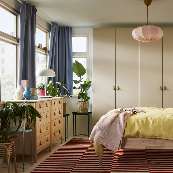 A bedroom with a bed frame and two chests of drawers in birch, a grey-beige wardrobe, blue curtains and yellow bed textiles.
