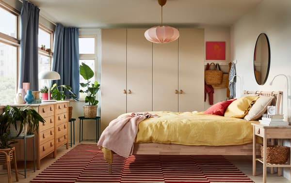 A bedroom with a bed frame and two chests of drawers in birch, a gray-beige wardrobe, blue curtains and yellow bed textiles.