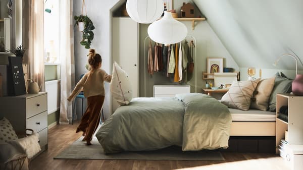 A bedroom with a girl running past a bed with grey striped bed linen and cushions, plus various storage and white pendants.