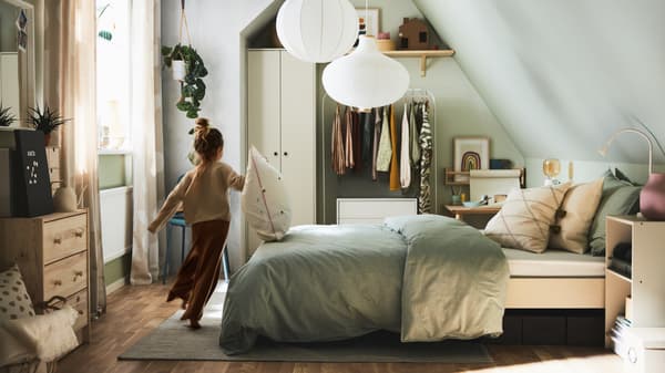 A bedroom with a girl running past a bed with grey striped bed linen and cushions, plus various storage and white pendants.