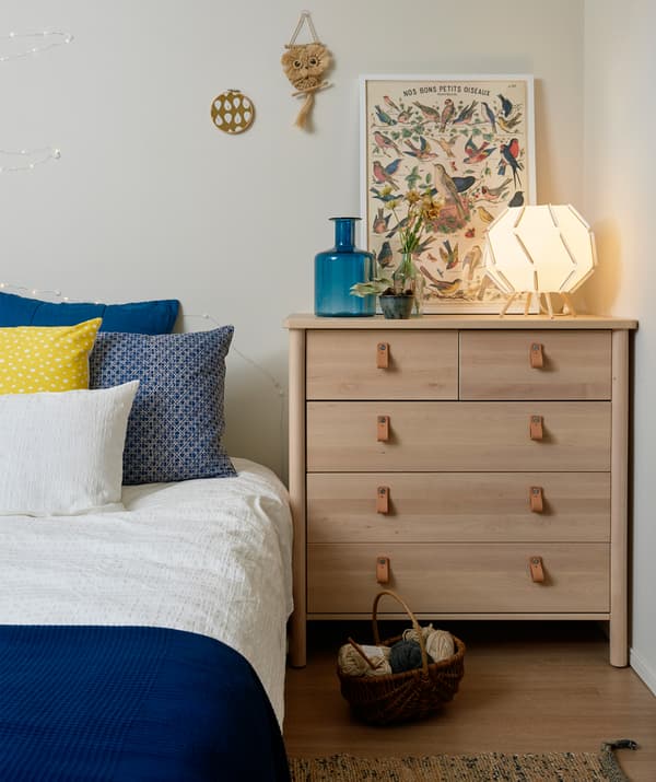 A bedroom with a pale wooden chest of drawers and bed dressed with blue and white bedding.