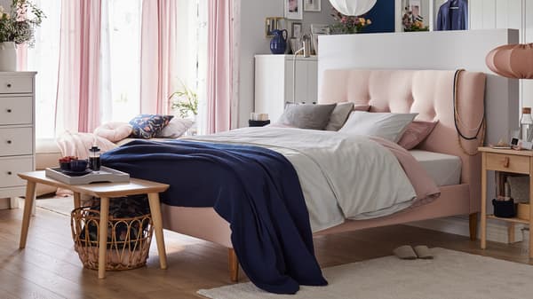 A bedroom with a pink bed with padded headboard, soft colored textiles, sheer pink curtains and wooden side tables.