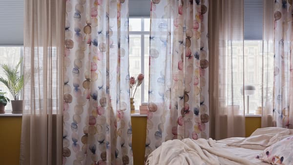 A bedroom with a row of windows with multicolour SÄCKMAL curtains, beige sheer curtains and white block-out cellular blinds.