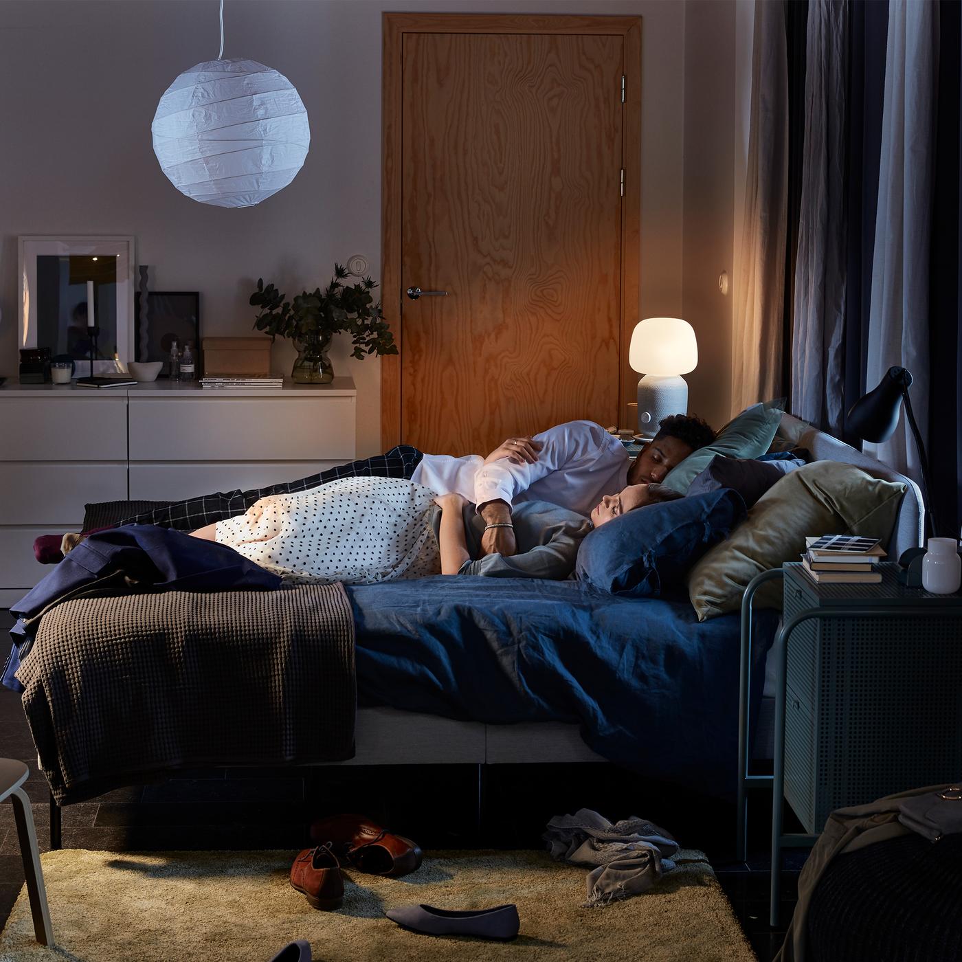 A bedroom with a SLATTUM upholstered bed frame, two lamps and dark blue quilt covers, and a couple sleeping on the bed.