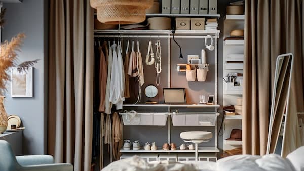 A bedroom with a small office occupying a few shelves of a storage system inside an open wardrobe behind curtains.