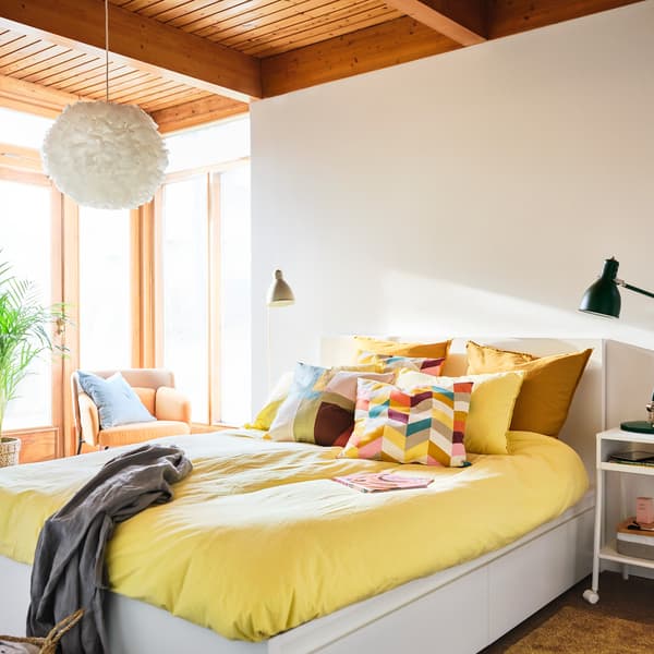 A bedroom with a white MALM bed frame, yellow quilt covers and cushions, and a HANNELISE cushion with a colorful pattern.