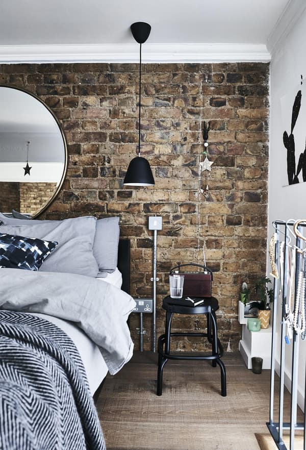 A bedroom with brick wall and grey bedding and a black stool being used as a bedside table.