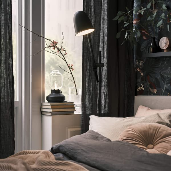 A bedroom with dark curtains hanging and a black lamp shining down on the bed.