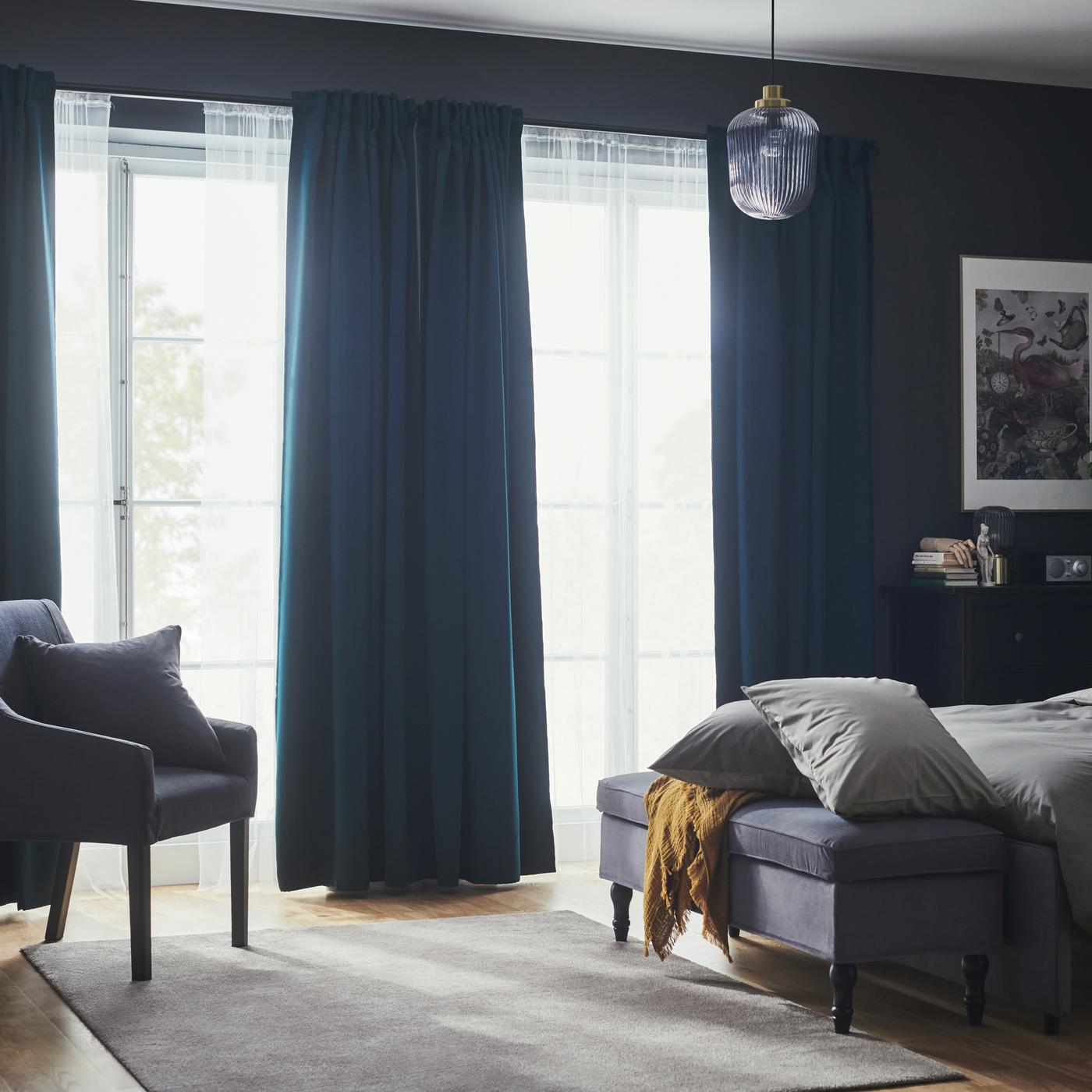 A bedroom with dark turquoise MAJGULL block-out curtains and net curtains in large windows near a bed and a bench.