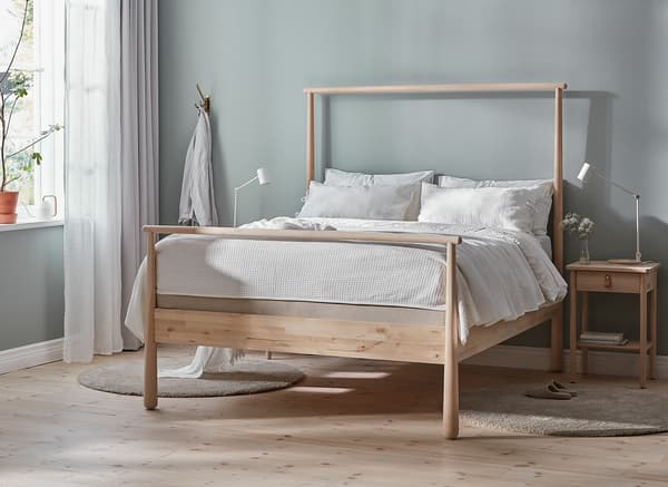 A bedroom with double, wooden bed frame, neutral bedding and a plant in the window.