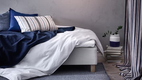 A bedroom with grey walls and a messy bed with an ÄNGSLILJA quilt cover, INDIRA bedspread in dark blue and three cushions.