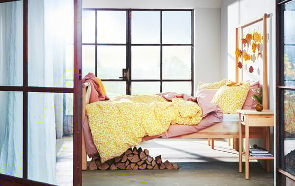 A bedroom with large windows, leaves hanging from a wooden bedframe and yellow and pink layered bedding.
