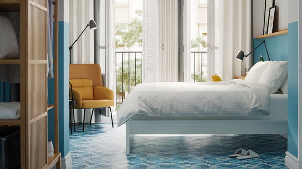 A bedroom with NORDKISA wardrobe in bamboo, a white bed with white bedlinen, blue walls and floors and a yellow armchair.