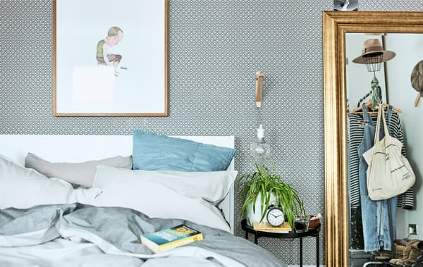 A bedroom with patterned wallpaper, bedding in shades of blue and a gold mirror reflecting a clothes rail.