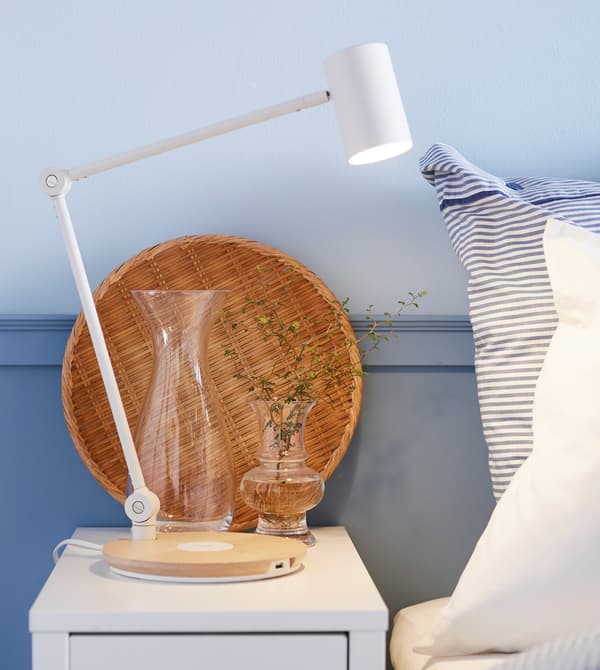 A bedside table is set with a lamp, vase of small flowers, and a wicker tray.
