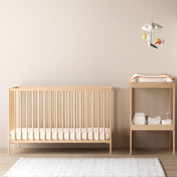 A beech SNIGLAR crib with only a mattress and fitted sheet in it, while a changing table is next to it with a  GULLIGAST mobile that hangs high above it.