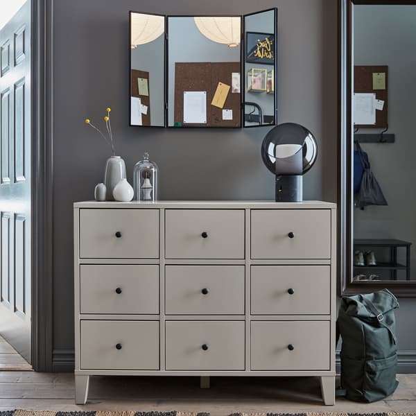 A beige BRYGGJA chest of drawers, a decorative table lamp, a full-body mirror, a wall-mounted mirror and a green backpack.