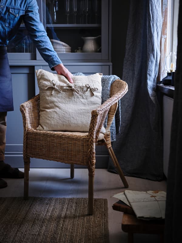 A beige-coloured AINA pillow is placed on a rattan AGEN chair that stands inside a dark blue living room.