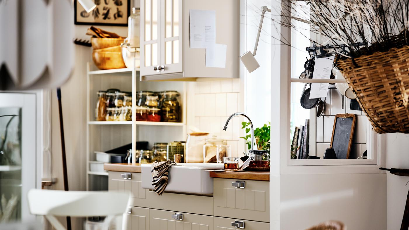 A beige country-style kitchen with STENSUND fronts on METOD cabinets, a white sink and shelving with jars and utensils.