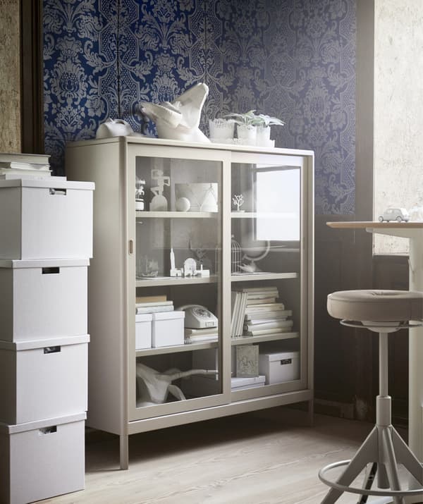 A beige glass-fronted cabinet in the corner of an office space with blue patterned wallpaper.