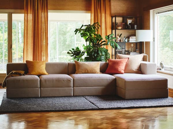 A beige JÄTTEBO corduroy corner sofa on a blue carpet in a living room with parquet flooring and yellow curtains. 