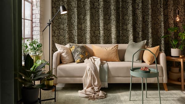 A beige LINANÄS sofa with various cushions on a light grey-green rug, in front of leaf patterned curtains hanging on a wall.