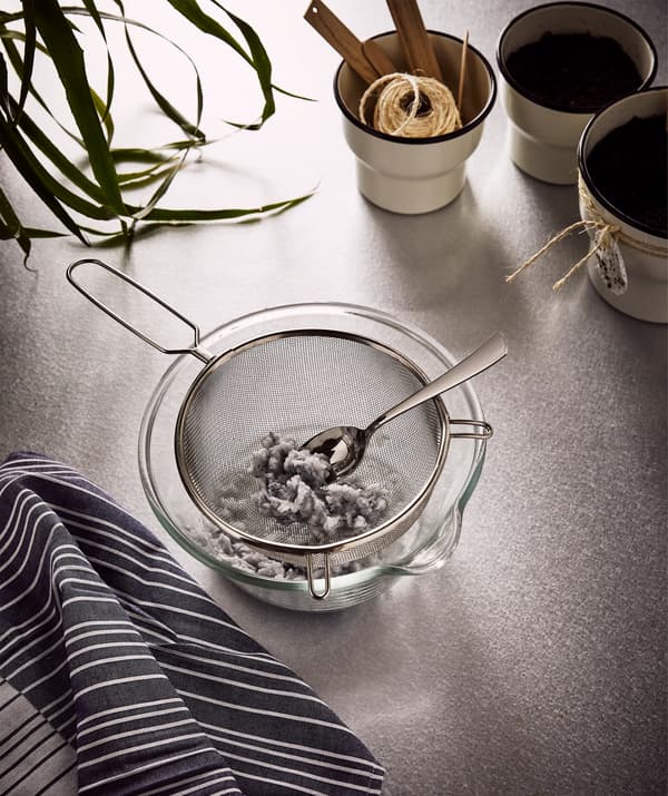 A big glass bowl with an IDEALISK strainer containing moist paper pulp from shredded napkins, beside three plant pots.