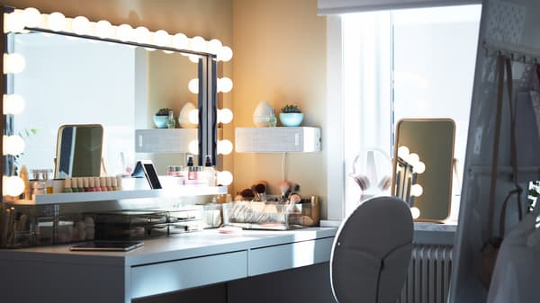 A big rectangular mirror with wall lamps above a white desk with lots of make-up storage, plus a white speaker on the wall.