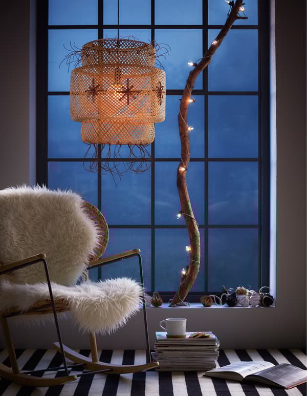 A big window with a hanging bamboo lamp decorated with holiday ribbon and a large branch standing on the sill with lights strung around it.