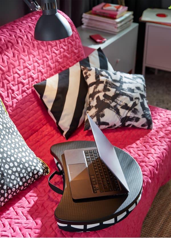 A black and white cushioned IKEA BYLLAN laptop support on a pink sofa amongst grey, black and white cushions.