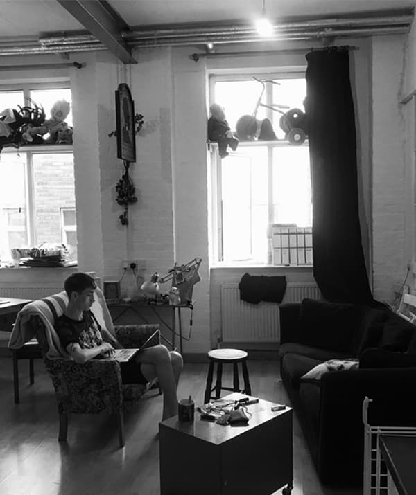 A black and white image of Ben sitting on a small sofa in the living area looking at his computer.