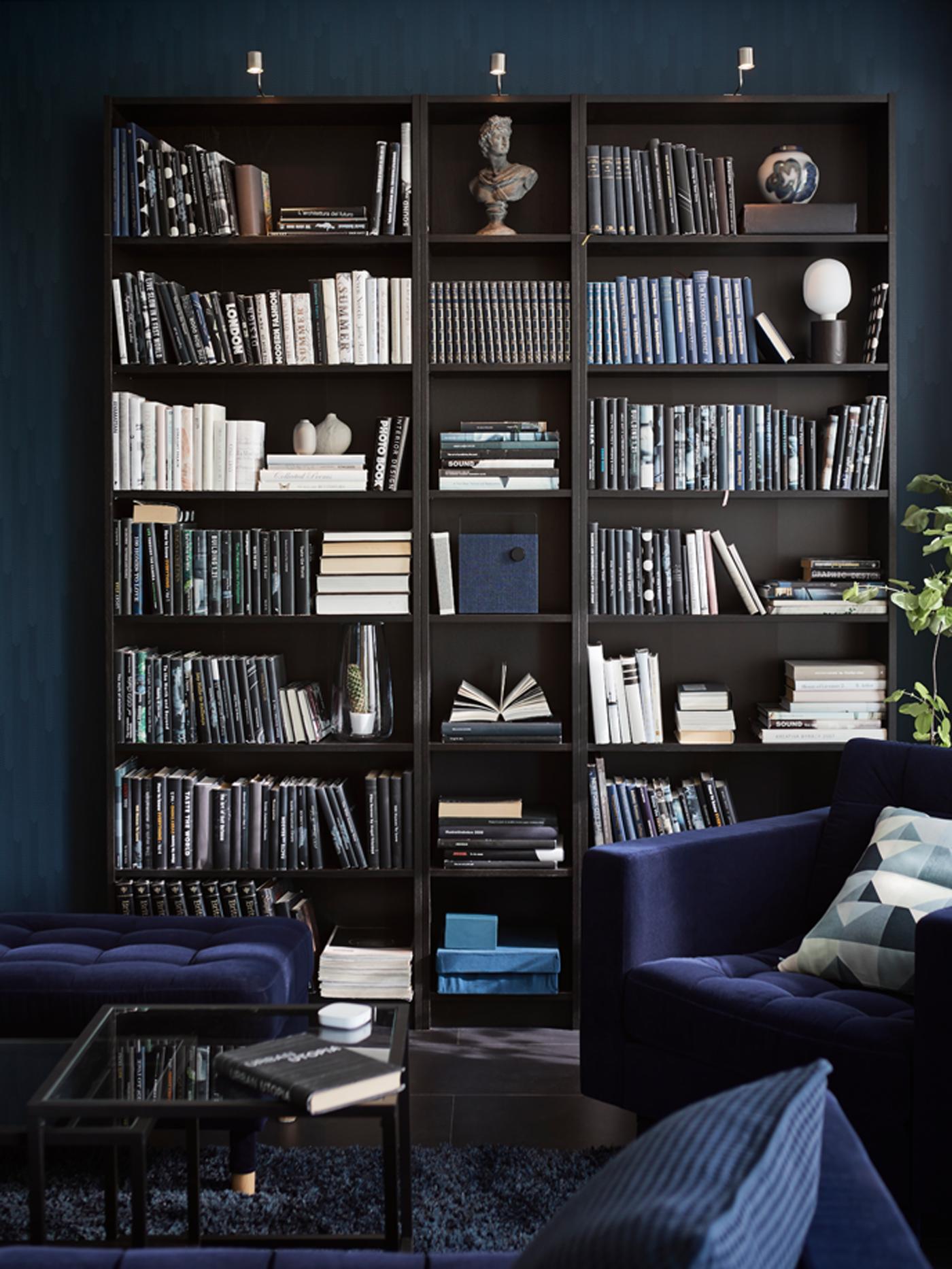 A black-brown BILLY bookcase in a blue living room filled with lots of books and a few objects. A blue armchair is in front.