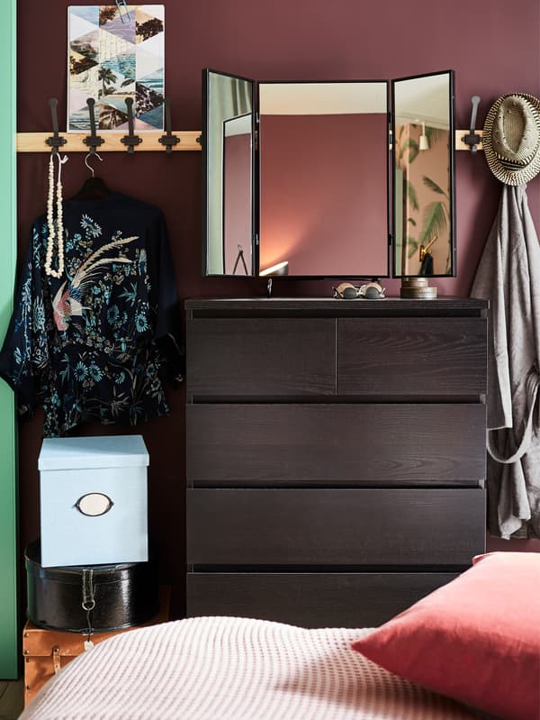A black-brown MALM chest of drawers, a black mirror, pine racks with grey hooks and a blue storage box with lid.