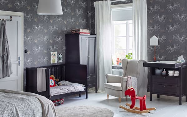 A black brown SUNDVIK cot and nappy changing table in the corner of a large adult bedroom with grey patterned wallpaper.