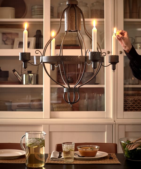 A black chandelier with four candles that are being lit by a person, a white glass-door cabinet and dinner served on a table.