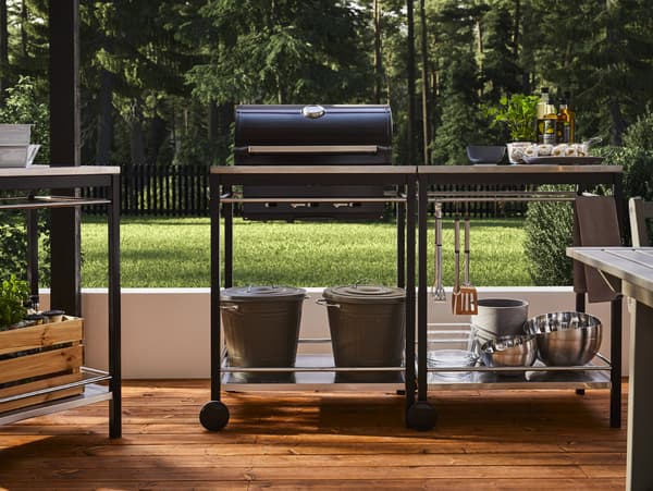 A black charcoal barbecue with trolley in stainless steel with kitchen utensils, bowls, bins with lids, herbs and oils.