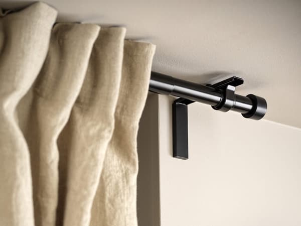 A black curtain rod with cream-coloured curtains pleated and hanging on it.