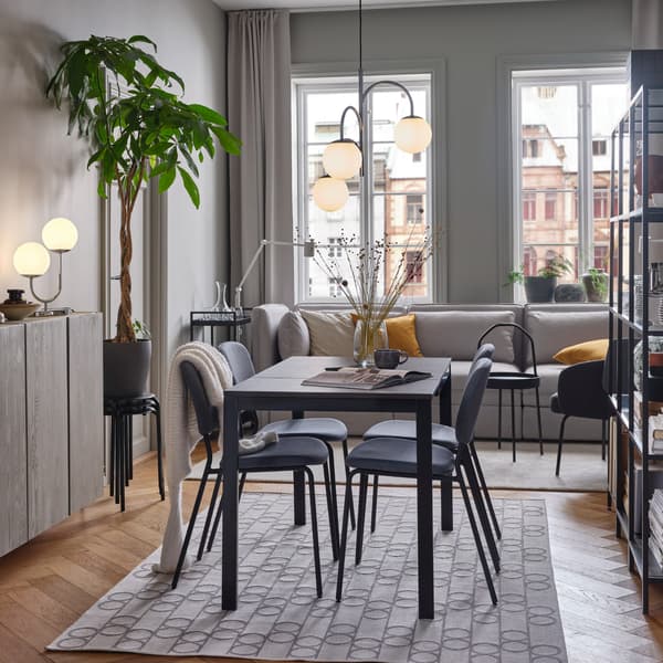 A black/dark brown table and grey chairs stand on a geometric-patterned rug. Above is a 3-armed chrome-plated pendant lamp.