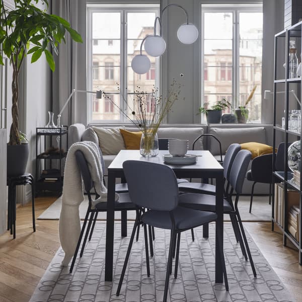 A black/dark brown table, grey chairs, a geometric-patterned rug, a white/chrome-plated pendant lamp and a light grey sofa.