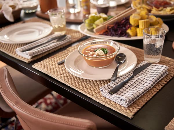 A black dining table with place mats made from water hyacinth, white plates, glass bowls with soup and folded tea towels.