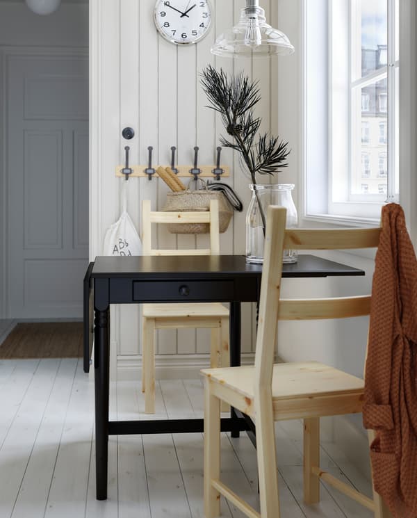 A black drop-leaf table with drawer, two pine chairs, a wooden rack with hooks, a wall clock and a glass pendant lamp.
