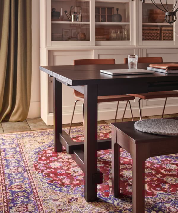 A black extendable table, two golden-brown leather chairs, a dark brown bench, a green curtain and an oriental-patterned rug.
