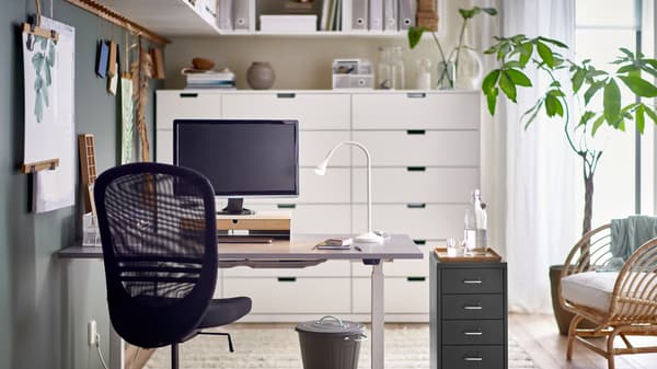 A black FLINTAN office chair and HELMER drawer unit by a grey RODULF sit/stand desk with a work lamp and computer screen.