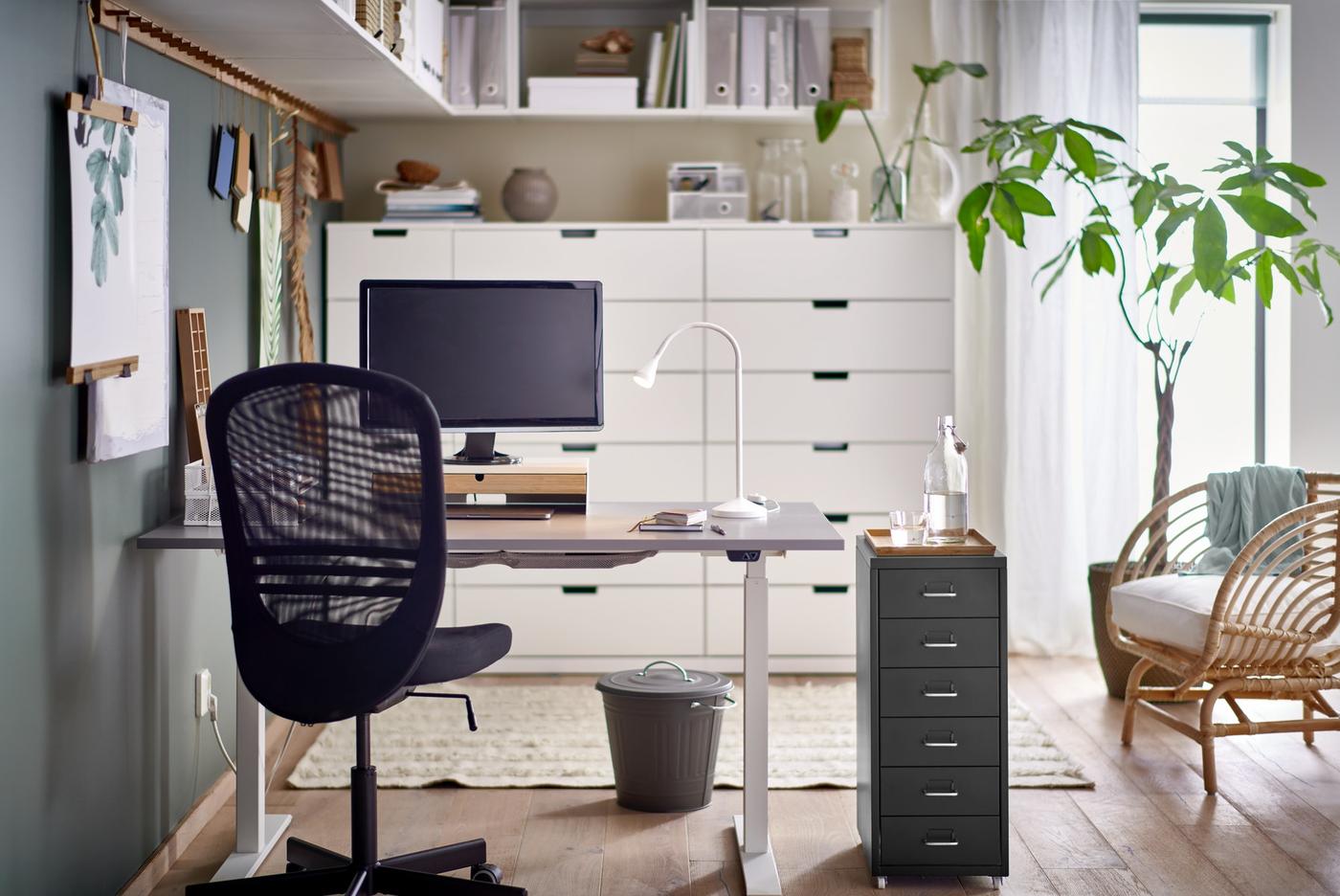 A black FLINTAN office chair and HELMER drawer unit by a grey RODULF sit/stand desk with a work lamp and computer screen.