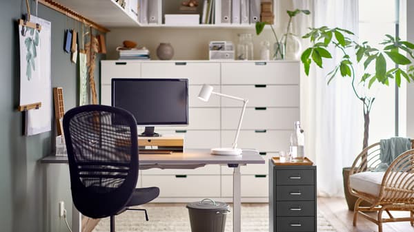 A black FLINTAN office chair and HELMER drawer unit by a grey RODULF sit/stand desk with a work lamp and computer screen.