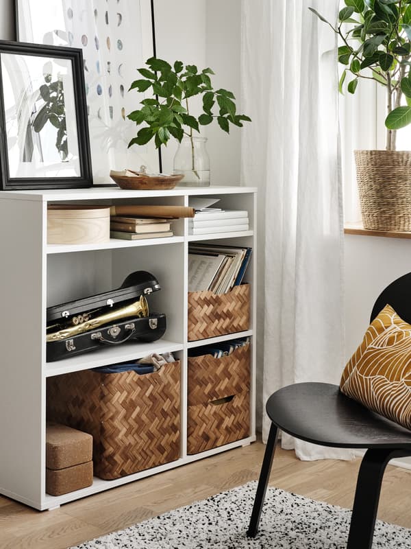 A black FRÖSET chair with a dark yellow cushion on it beside an open shelving unit holding baskets, books and a trumpet.