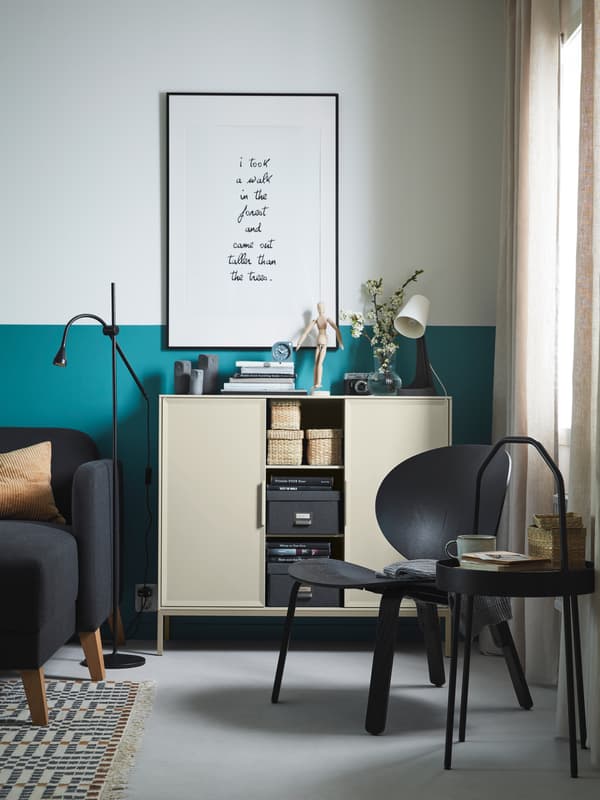 A black FRÖSET easy chair beside a curtained-window with a round black side table beside it holding a cup and a small basket.